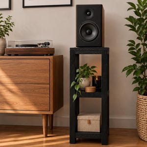 Op de afbeelding: Een hoge, zwarte houten standaard met drie open planken. De bovenste plank bevat een zwarte luidspreker. De middelste plank bevat een kleine groene plant in een pot, een donkere glazen kaars en houten onderzetters.