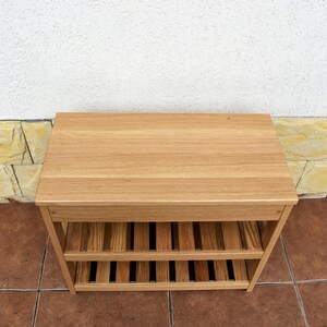 May include: A rectangular light brown wooden shoe rack featuring a solid flat top surface and two lower slatted shelves. The unit is constructed from natural wood with a visible grain pattern, providing open storage space for footwear or other items.