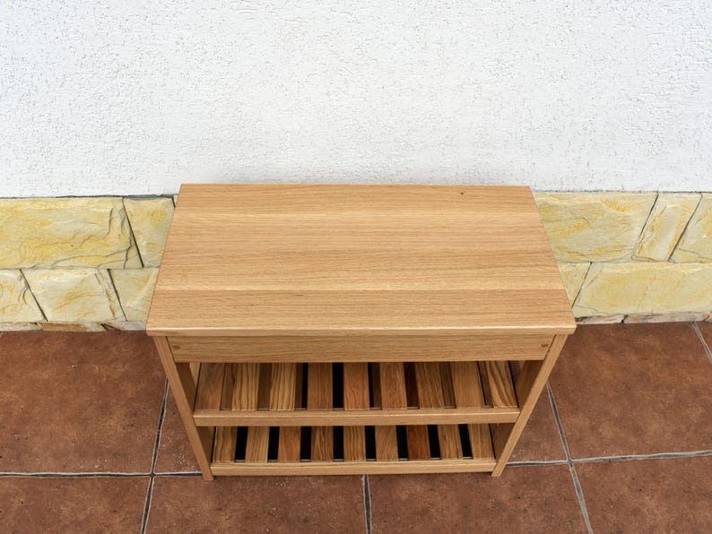 May include: A rectangular light brown wooden shoe rack featuring a solid flat top surface and two lower slatted shelves. The unit is constructed from natural wood with a visible grain pattern, providing open storage space for footwear or other items.