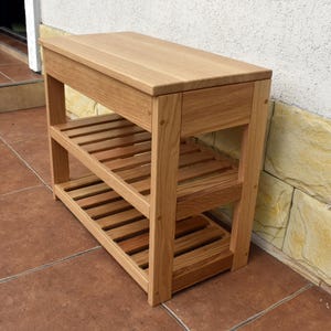 May include: A light brown wooden console table featuring a solid rectangular top surface and two lower slatted shelves. The structure is supported by four vertical legs, showcasing a natural wood grain finish throughout the entire piece of furniture.