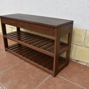 May include: A rectangular dark brown wooden bench featuring a solid flat top surface and two lower slatted shelves. The furniture piece is constructed with four sturdy vertical legs and horizontal support beams, showcasing a visible wood grain texture.