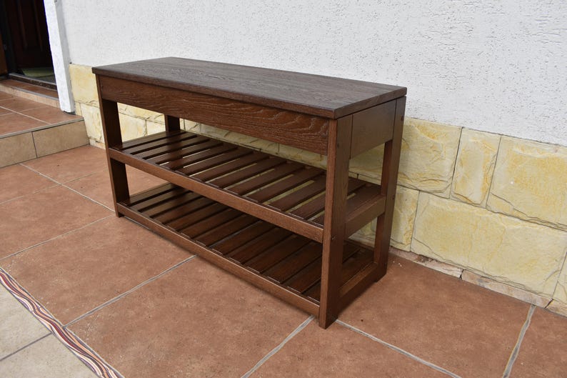 May include: A rectangular dark brown wooden bench featuring a solid flat top surface and two lower slatted shelves. The furniture piece is constructed with four sturdy vertical legs and horizontal support beams, showcasing a visible wood grain texture.