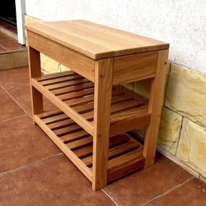 May include: A rectangular wooden shoe rack with a smooth top surface and two lower slatted shelves. The unit is constructed from light brown natural wood with visible grain patterns, featuring four sturdy vertical legs and a clean, open design.