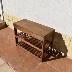May include: A rectangular wooden bench with a smooth top surface and two lower slatted shelves. The furniture piece features a medium brown finish and is constructed with four vertical legs supporting the structure in a simple, functional design.