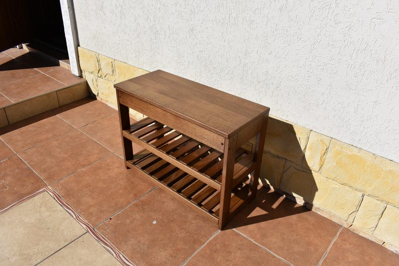 May include: A rectangular wooden bench with a smooth top surface and two lower slatted shelves. The furniture piece features a medium brown finish and is constructed with four vertical legs supporting the structure in a simple, functional design.