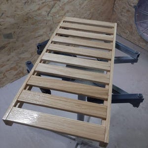 May include: A rectangular wooden slatted rack featuring a light brown natural wood grain finish. The structure consists of evenly spaced horizontal wooden planks secured by two side rails, creating a grid-like surface with a smooth, glossy texture.