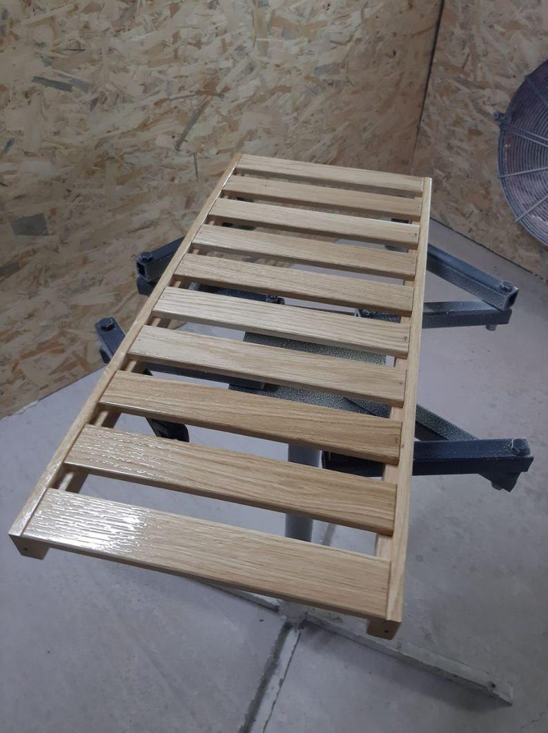 May include: A rectangular wooden slatted rack featuring a light brown natural wood grain finish. The structure consists of evenly spaced horizontal wooden planks secured by two side rails, creating a grid-like surface with a smooth, glossy texture.