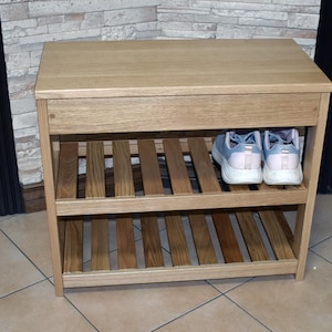 May include: A light brown wooden shoe rack featuring a solid top surface and two slatted shelves for storage. The unit has a rectangular frame with vertical side supports. A pair of light blue and white athletic sneakers sits on the top shelf.
