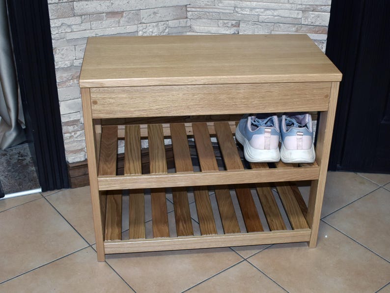 May include: A light brown wooden shoe rack featuring a solid top surface and two slatted shelves for storage. The unit has a rectangular frame with vertical side supports. A pair of light blue and white athletic sneakers sits on the top shelf.