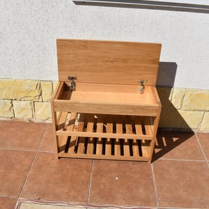 May include: A light brown wooden storage bench featuring a hinged lid that opens to reveal a hollow compartment. The unit includes two slatted shelves underneath the main storage area, constructed from light-toned wood with visible grain patterns.
