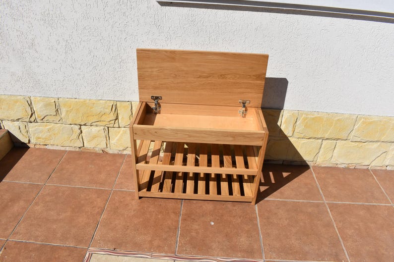 May include: A light brown wooden storage bench featuring a hinged lid that opens to reveal a hollow compartment. The unit includes two slatted shelves underneath the main storage area, constructed from light-toned wood with visible grain patterns.