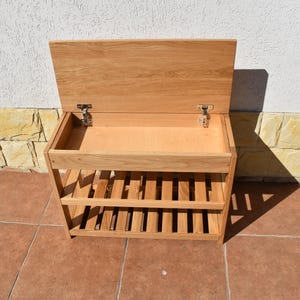 May include: A light brown wooden storage bench featuring a hinged lid that opens to reveal a hollow compartment. Below the storage area are two slatted shelves designed for organization. The piece is constructed from natural wood with visible grain.