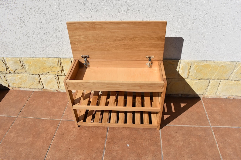 May include: A light brown wooden storage bench featuring a hinged lid that opens to reveal a hollow compartment. Below the storage area are two slatted shelves designed for organization. The piece is constructed from natural wood with visible grain.