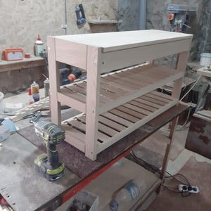 May include: A light-colored wooden shoe rack featuring a solid top surface and two slatted shelves below. The structure is held together by visible metal screws on the side panels, showcasing a natural wood grain finish and a rectangular frame.