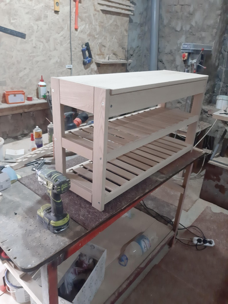 May include: A light-colored wooden shoe rack featuring a solid top surface and two slatted shelves below. The structure is held together by visible metal screws on the side panels, showcasing a natural wood grain finish and a rectangular frame.