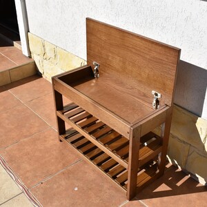 May include: A brown wooden storage bench with a hinged lid that opens to reveal a hollow compartment. The unit features two lower slatted shelves for storage and is constructed with visible wood grain, metal hinges, and a rectangular frame.