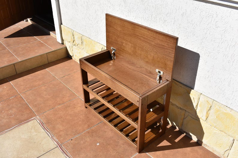 May include: A brown wooden storage bench with a hinged lid that opens to reveal a hollow compartment. The unit features two lower slatted shelves for storage and is constructed with visible wood grain, metal hinges, and a rectangular frame.