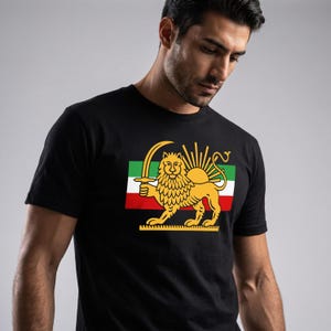 Puede incluir: Camiseta negra con un león dorado sosteniendo una espada, un sol y un diseño de bandera. La bandera tiene rayas verdes, blancas y rojas. El gráfico del león está centrado en la camiseta.