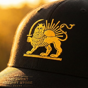 Puede incluir: Una gorra de béisbol negra con un emblema bordado en dorado de un león sosteniendo una espada frente a un sol naciente. El bordado tiene un acabado texturizado. El texto THE GREAT PATRIOT STORE es visible en la parte inferior de la imagen.