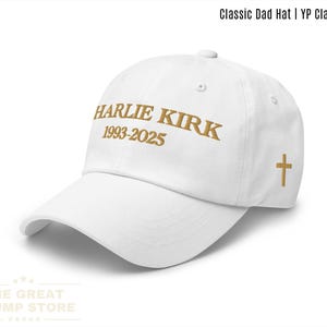 Puede incluir: Gorra blanca clásica con el texto "HARLIE KIRK 1993-2025" en letras doradas. Una cruz dorada está en el lateral. La gorra es de The Great Trump Store.