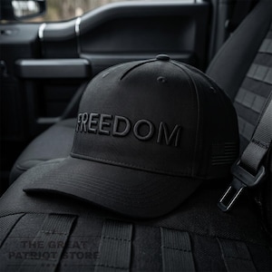 Könnte beinhalten: Schwarze Baseballkappe mit dem Wort "FREEDOM" auf der Vorderseite gestickt. Die Kappe hat auch ein kleines amerikanisches Flaggen-Design an der Seite. Die Kappe liegt auf einem schwarzen Sitz.