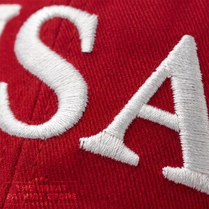 May include: A red fabric surface featuring large, white, embroidered letters that spell out USA. The stitching is dense and creates a textured, raised appearance against the vibrant red background of the material, which shows a clear woven pattern.