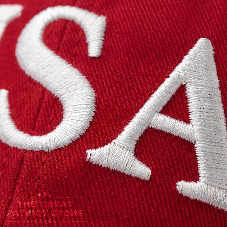 May include: A red fabric surface featuring large, white, embroidered letters that spell out USA. The stitching is dense and creates a textured, raised appearance against the vibrant red background of the material, which shows a clear woven pattern.