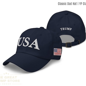 Puede incluir: Gorra clásica azul marino con "USA" bordado en blanco en la parte delantera y "TRUMP" en la parte trasera. Cuenta con un parche de la bandera estadounidense en el lateral. Correa ajustable. The Great Trump Store.