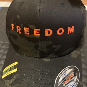 Könnte beinhalten: Eine schwarze Baseballkappe mit Tarnmuster und dem in orangefarbenem Garn auf die Vorderseite gestickten Wort FREEDOM. Die Kappe hat ein gelbes Multicam Black-Etikett und einen runden Flexfit-Aufkleber auf dem Schirm, der die Größe S/M angibt.
