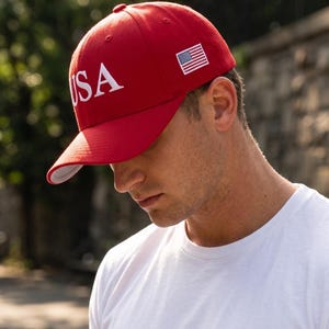 May include: A bright red baseball cap featuring the white embroidered text USA on the front panel and a small embroidered American flag patch on the side. The cap has a structured crown, a curved brim, and a white underside on the brim.