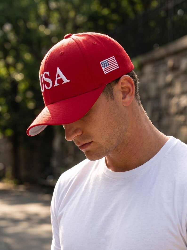 May include: A bright red baseball cap featuring the white embroidered text USA on the front panel and a small embroidered American flag patch on the side. The cap has a structured crown, a curved brim, and a white underside on the brim.