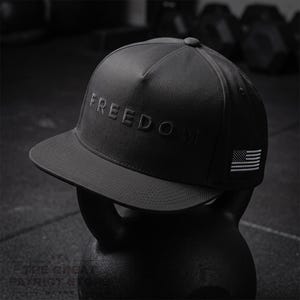 Könnte beinhalten: Eine schwarze Baseballkappe mit flachem Schirm und strukturierter Krone. Das Wort FREEDOM ist in Schwarz auf die Vorderseite geprägt. Ein kleiner, rechteckiger Aufnäher mit einer weiß gestickten amerikanischen Flagge befindet sich an der Seite.