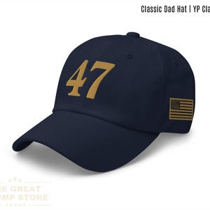 以下が含まれることがあります： ネイビーブルーの野球帽。前面に金色の数字「47」が刺繍されています。側面にはアメリカ国旗のパッチがあり、「Classic Dad Hat | YP Classics®」の文字があります。