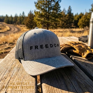 May include: A gray trucker hat with the word "FREEDOM" in black block letters. The hat has a mesh back and a curved brim. The hat is sitting on a wooden picnic table with a pair of work gloves and a thermos.
