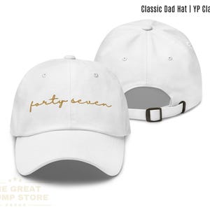 May include: White Classic Dad Hat with the text "forty seven" in gold script. The hat has a curved brim and an adjustable metal buckle closure. The hat is made of a soft, breathable material.