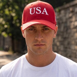 May include: A bright red baseball cap featuring the letters USA embroidered in white on the front panel. The cap has a structured crown, a curved brim, and a small American flag patch visible on the side panel. The design is simple and bold.