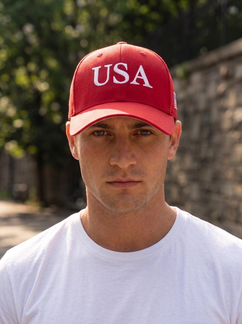 May include: A bright red baseball cap featuring the letters USA embroidered in white on the front panel. The cap has a structured crown, a curved brim, and a small American flag patch visible on the side panel. The design is simple and bold.
