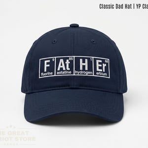 May include: A navy blue baseball cap featuring a white graphic design of the word FATHER spelled out using periodic table elements. Each letter block includes the chemical symbol, atomic number, and element name: Fluorine, Astatine, Hydrogen, Erbium.