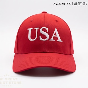May include: A bright red baseball cap featuring the text USA embroidered in white on the front. The cap is labelled as a Flexfit Wooly Combed Cap. The design includes a structured crown and a curved brim, presenting a clean and simple aesthetic.