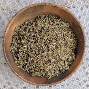 Peut inclure: Un bol en bois rempli d'herbes séchées. Les herbes sont un mélange de vert et de brun, avec un aspect texturé. Le bol est rond et en bois clair.