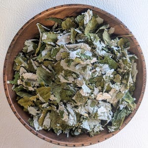 May include: A wooden bowl filled with dried green and white leaves. The leaves have a textured appearance and are arranged in a circular pattern within the bowl. The bowl is made of a dark wood.