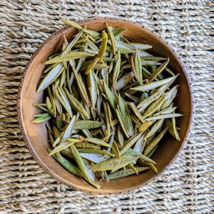 May include: A round wooden bowl filled with a collection of dried, elongated green leaves. The leaves are narrow, pointed, and display various shades of olive and sage green, with some showing lighter, silvery-toned undersides within the bowl.
