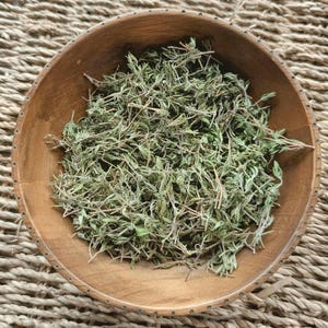 May include: A round, medium-brown wooden bowl filled with a generous portion of dried green thyme leaves and stems. The bowl features a smooth, polished interior surface and a simple, circular rim, containing the loose, aromatic dried herb mixture.