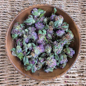 May include: A round, dark brown wooden bowl filled with a collection of dried purple and green clover flowers. The dried blossoms exhibit varied shades of violet, lavender, and muted green foliage, clustered together within the smooth, circular wooden vessel.