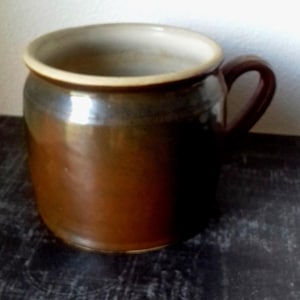 Peut inclure: Une tasse en céramique brune avec un bord brun clair et une anse. La tasse est posée sur une surface noire avec un motif blanc.