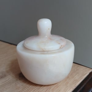 Peut inclure: Un petit pot en pierre blanc cassé, de forme ronde, avec une finition lisse et polie et de légères veines beiges. Le pot est doté d'un couvercle amovible assorti avec une poignée arrondie en forme de bouton sur le dessus.
