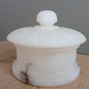 May include: A white, lidded jar made of a translucent material, possibly alabaster or marble. The jar has a rounded base and a domed lid with a small knob. The surface has subtle veining and a few brown streaks.