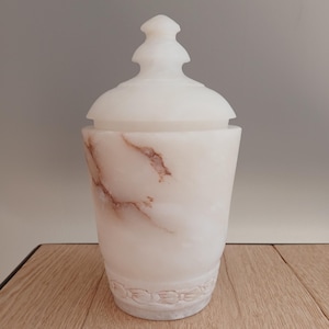Puede incluir: Un tarro cilíndrico de mármol blanco con tapa extraíble que presenta un remate puntiagudo. La piedra muestra vetas naturales de color marrón claro por todo el cuerpo. Una banda decorativa tallada con un motivo de lazos rodea la base.