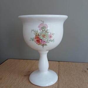 Puede incluir: Cuenco de pedestal de cerámica blanca con diseño floral. El cuenco tiene un borde ancho y acampanado y un cuerpo redondeado, sostenido por una base de pedestal. El diseño floral presenta flores rosas, melocotón y verdes. El cuenco probablemente se utiliza con fines decorativos.