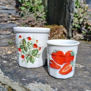 Peut inclure: Deux récipients en céramique blanche décorés de fraises. L'un présente un motif de fraisier avec des baies rouges et des feuilles vertes. L'autre montre des fraises rouges en gros plan. Les deux sont sur une surface en pierre.